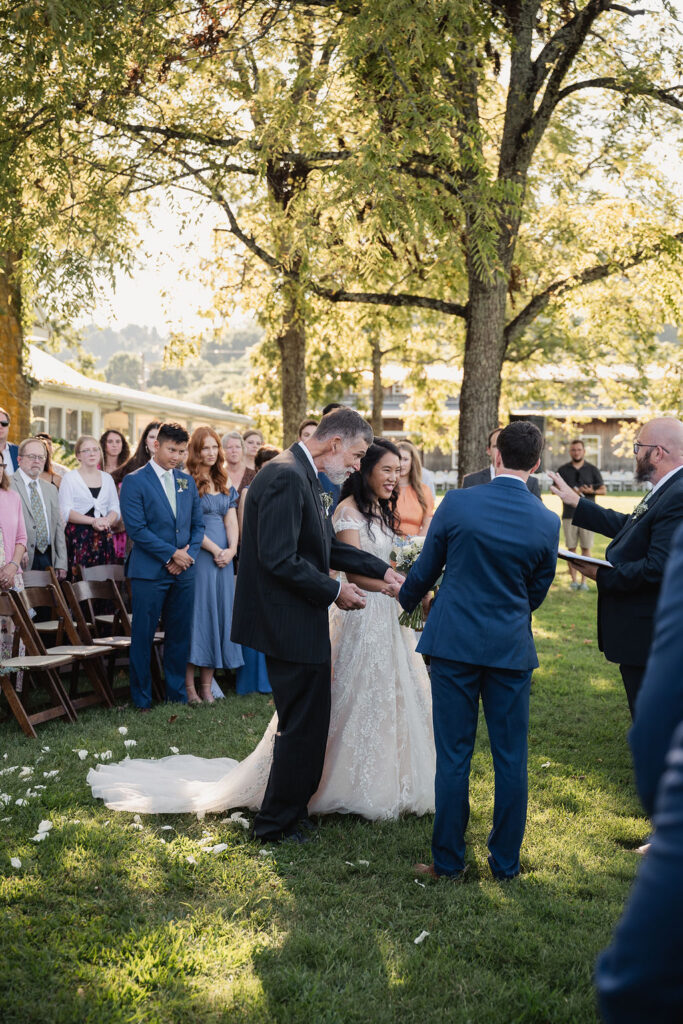 S and her father meet L at the lakeside altar, literally surrounded by hundreds of friends, and the organization of chairs softens the crowd to make a large wedding feel intimate.
